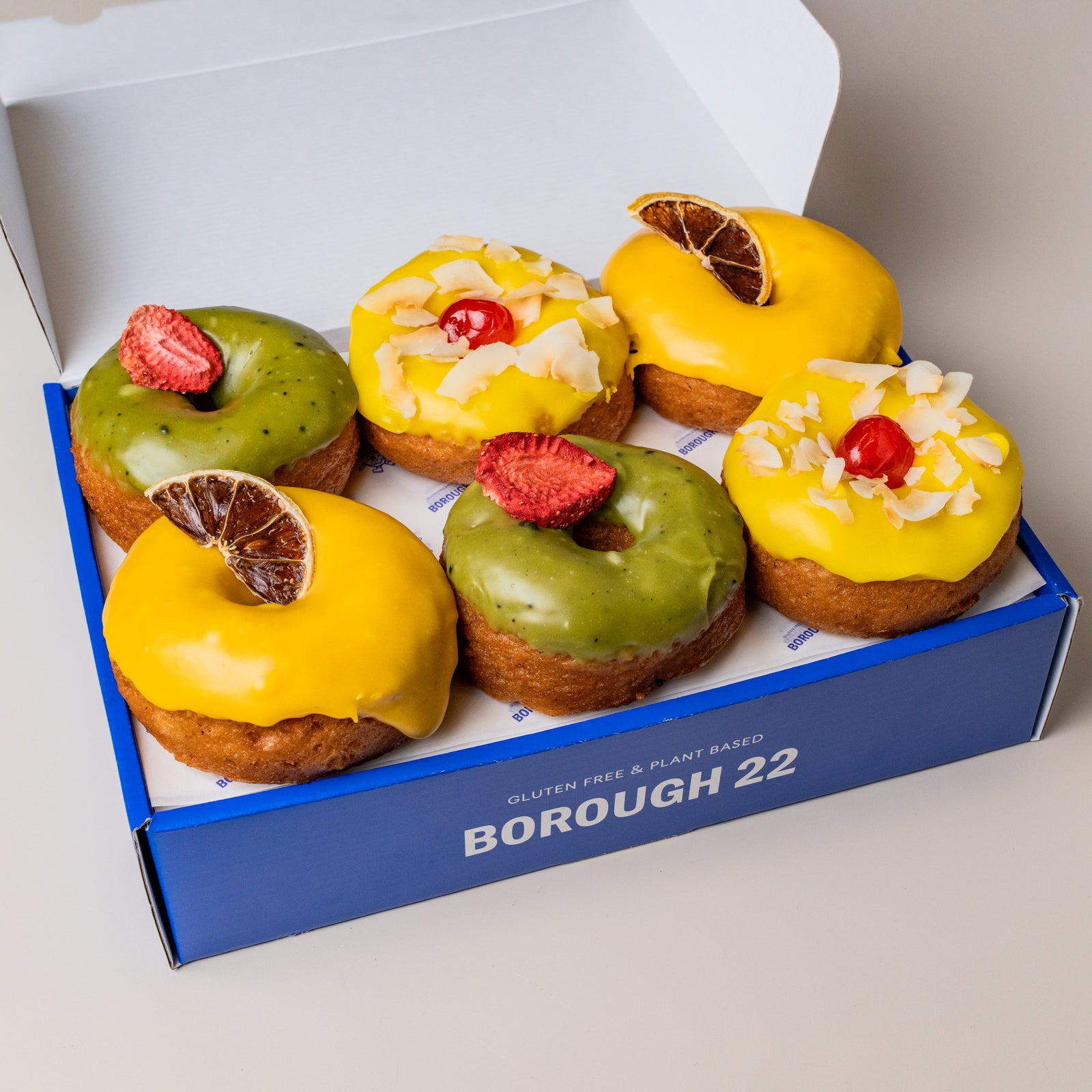 Six decorated gluten-free and plant-based donuts in a blue Borough 22 box. The donuts have yellow and green glazes, topped with strawberries, coconut flakes, red cherries, and dried orange slices.