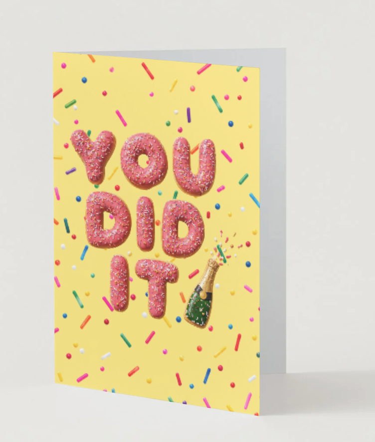 A yellow greeting card with the words 'YOU DID IT' spelled out in pink sprinkle-text letters, decorated with multicolored sprinkles and a small champagne bottle illustration, shown standing against a light gray background.