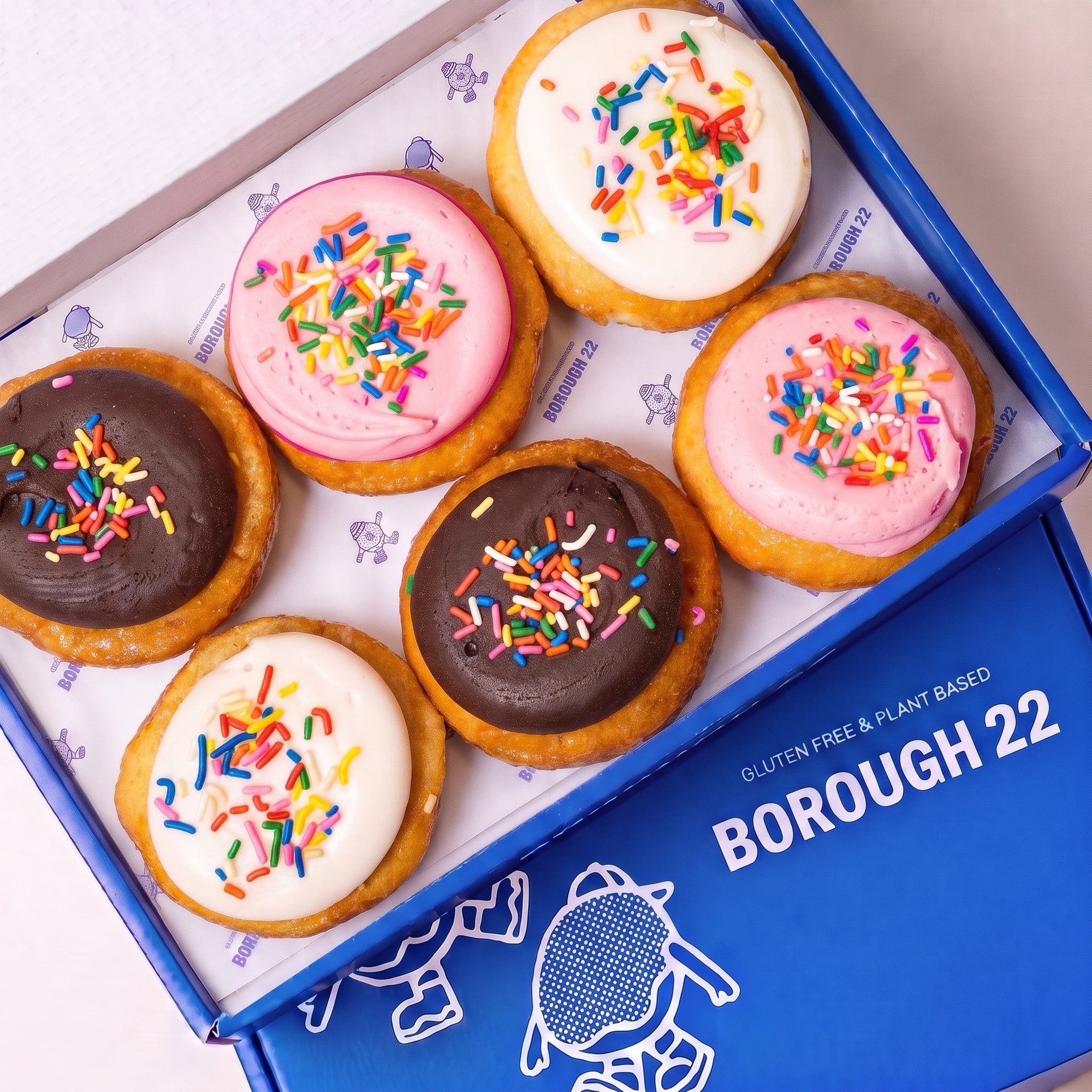 Open Borough 22 box of six gluten free vegan baked doughnuts iced in pink, white and chocolate, each covered in rainbow sprinkles