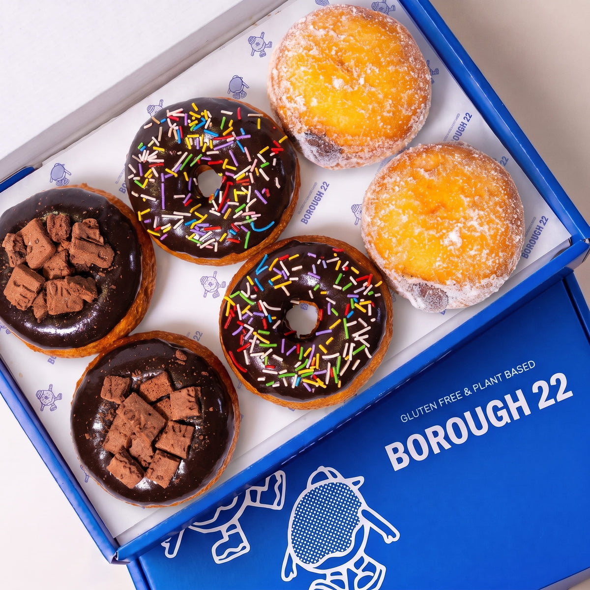 Open Borough 22 box of six gluten free oat free vegan fried chocolate doughnuts, chocolate iced rings with rainbow sprinkles, chocolate iced topped with chocolate biscuit pieces, and sugared filled rounds