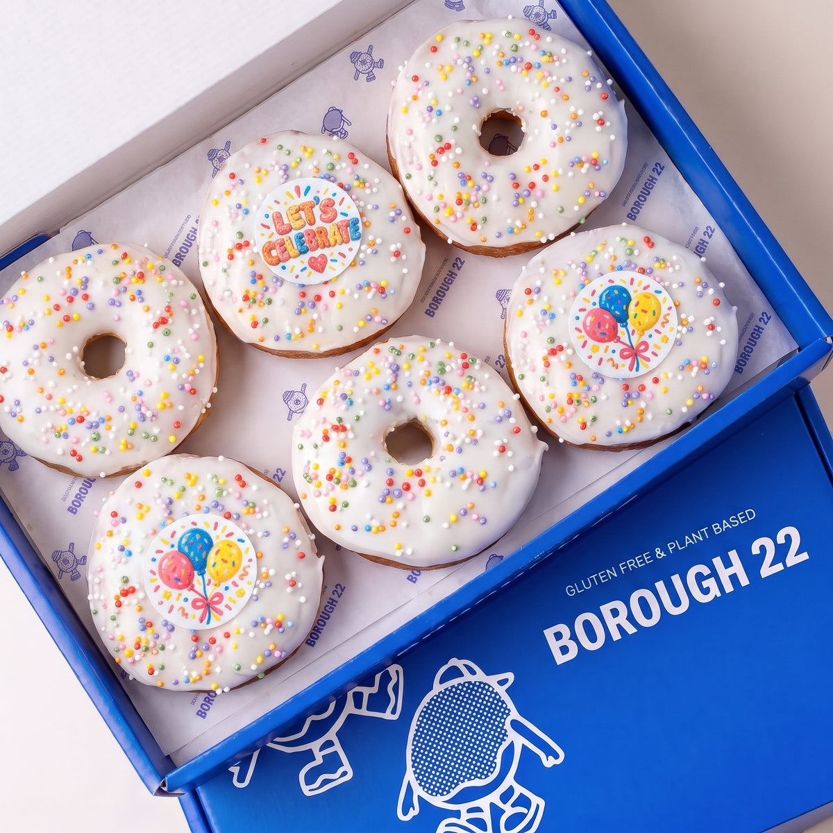 Gluten free vegan party doughnuts with white icing, rainbow sprinkles and Let's Celebrate and balloon toppers in a Borough 22 box