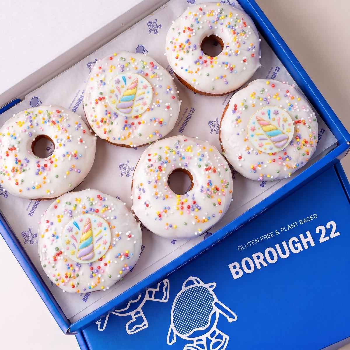 Gluten free vegan unicorn doughnuts with white icing, pastel sprinkles and rainbow unicorn horn toppers in a Borough 22 box