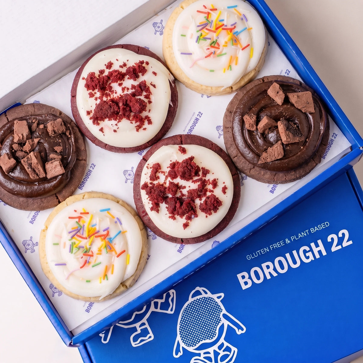 Six gluten free cookies in a blue Borough 22 box: two red velvet with white icing and red crumb, two funfetti with white icing and rainbow sprinkles, and two double chocolate crunch with chocolate icing and chocolate chunks.