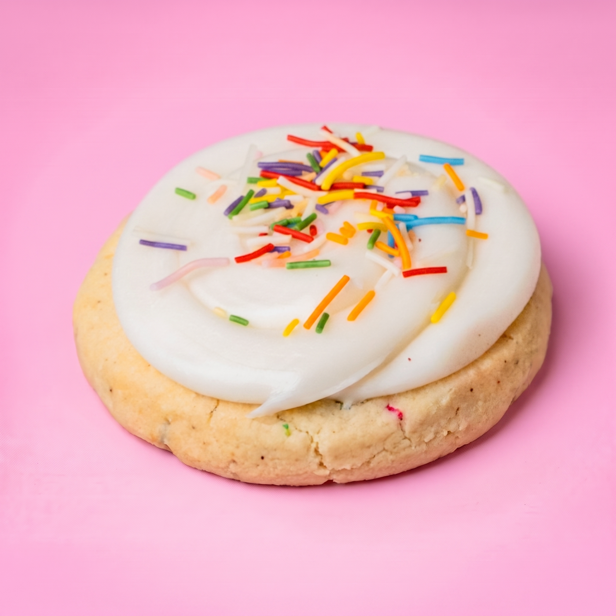 A round sugar cookie topped with thick white frosting and colorful sprinkles, photographed on a solid pink background.