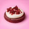 A red velvet gluten-free cookie with a thick swirl of white frosting and sprinkled with red cake crumbs, set against a solid pink background.